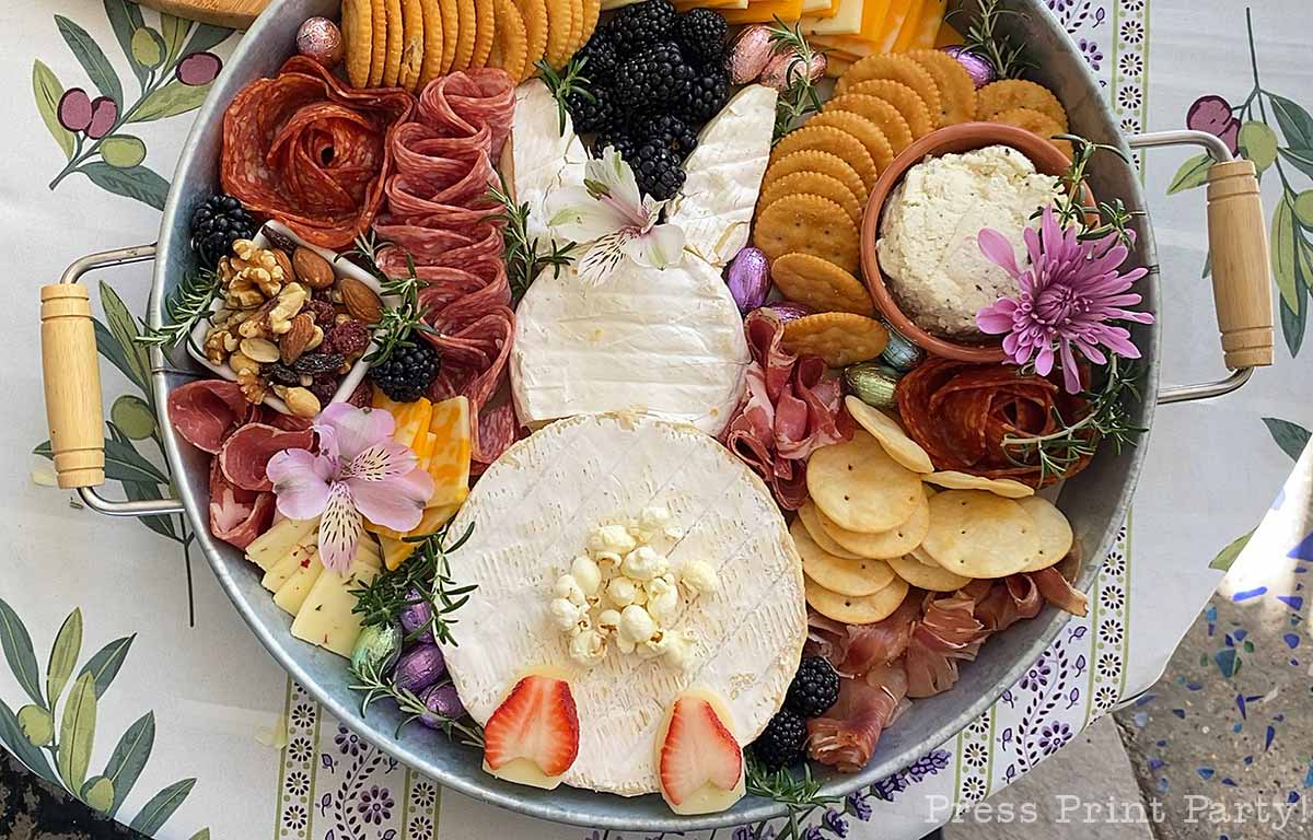 A round Easter Bunny Charcuterie Board featuring brie, sliced meats, cheeses, crackers, blackberries, nuts, flowers, and herbs is artfully arranged on a table with a floral tablecloth.