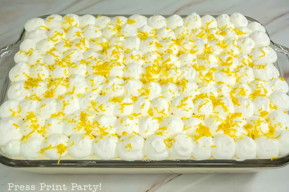 A rectangular limoncello tiramisu topped with piped whipped cream and sprinkled with lemon zest sits in a glass baking dish on a marble surface.
