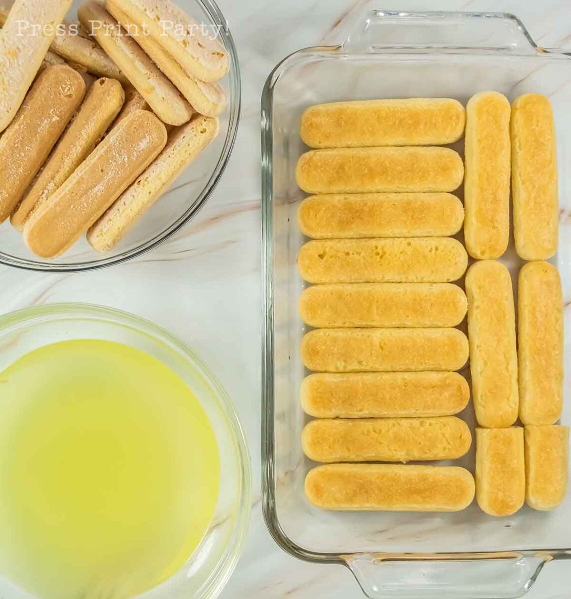 Overhead view of ladyfingers in a glass baking dish, a bowl of plain ladyfingers, and a bowl of yellow liquid on a marble surface, all set for preparing limoncello tiramisu.