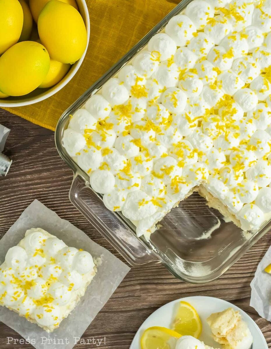 A glass baking dish of limoncello tiramisu topped with whipped cream and lemon zest, with one square served on parchment nearby. Fresh lemons and lemon wedges are beside the dessert on a wooden table.