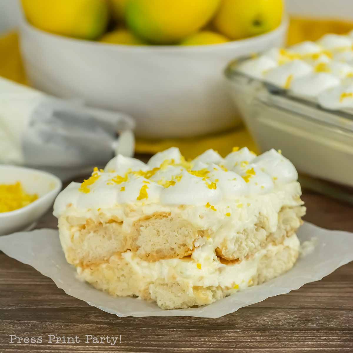 A slice of creamy limoncello tiramisu with layers of cake and whipped topping sits on parchment paper, garnished with lemon zest. A bowl of lemons and a pan of the same dessert are in the background.