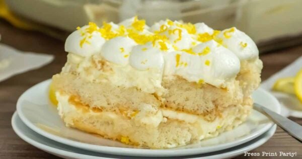 A close-up of a slice of limoncello tiramisu on a white plate, topped with whipped cream and lemon zest, showing creamy layers and a fork on the side.