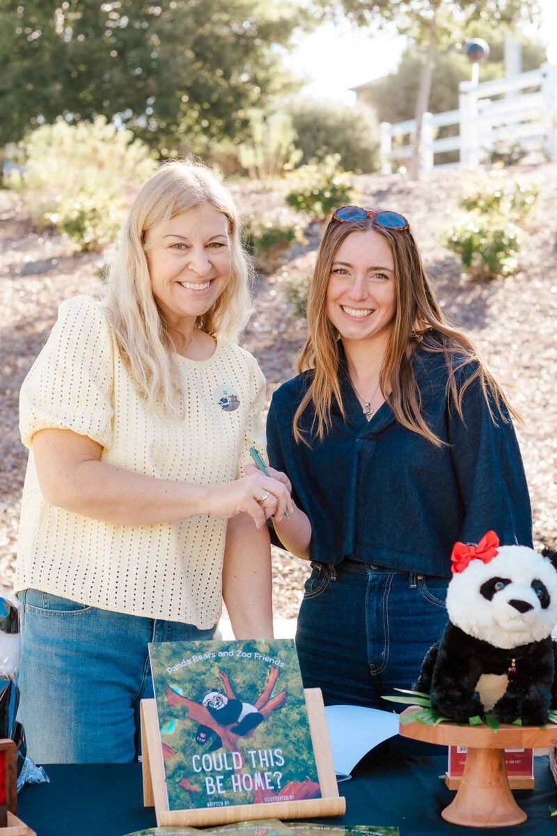 Two women smile at an outdoor table displaying a children’s book titled “Could This Be Home?” and a stuffed panda, perfect for sparking panda party ideas. Trees and a white fence are in the background on a sunny day.
