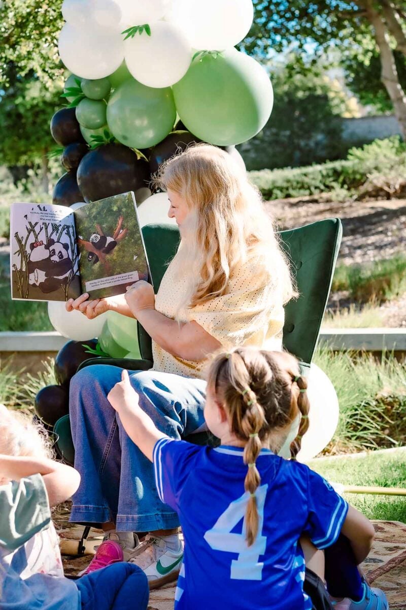 A woman with long blonde hair reads a picture book featuring a panda with balloons to a group of young children outdoors, near green and white balloons, with trees and sunlight in the background.