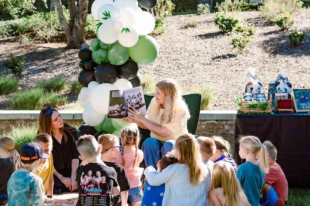 A woman reads a picture book to young children seated outdoors, while another adult sits nearby. Behind them, decorative balloons—including one shaped like a panda with balloons—frame a table with gift bags and books on this sunny day.