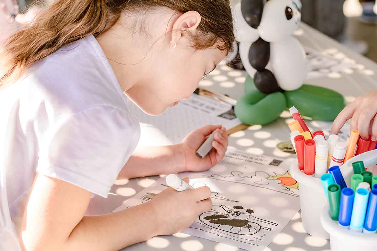 A young girl colors a picture of a panda at a table covered with markers and art supplies—perfect inspiration for panda party ideas. A panda-shaped balloon sits among the papers, as sunlight creates dappled patterns on her and the table.