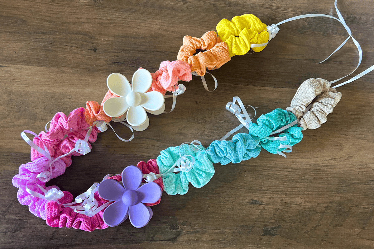 A colorful lei made of fabric scrunchies in pink, orange, yellow, green, and teal with two large plastic flowers and white ribbon accentsβperfect for unique graduation leis ideasβdisplayed on a wooden surface.