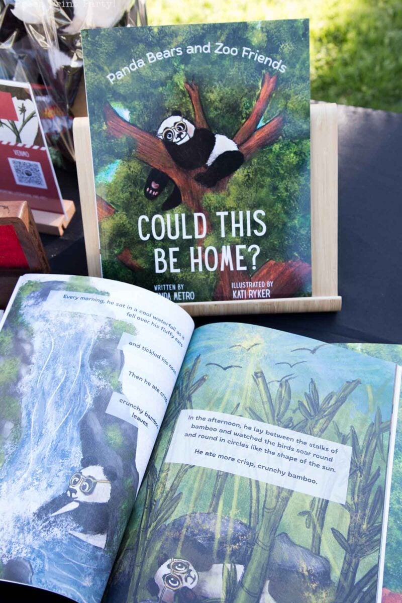 A children’s book titled “Could This Be Home?” featuring a panda bear on the cover is displayed on a wooden stand, with the book open to illustrated pages showing a panda with balloons in a bamboo forest.