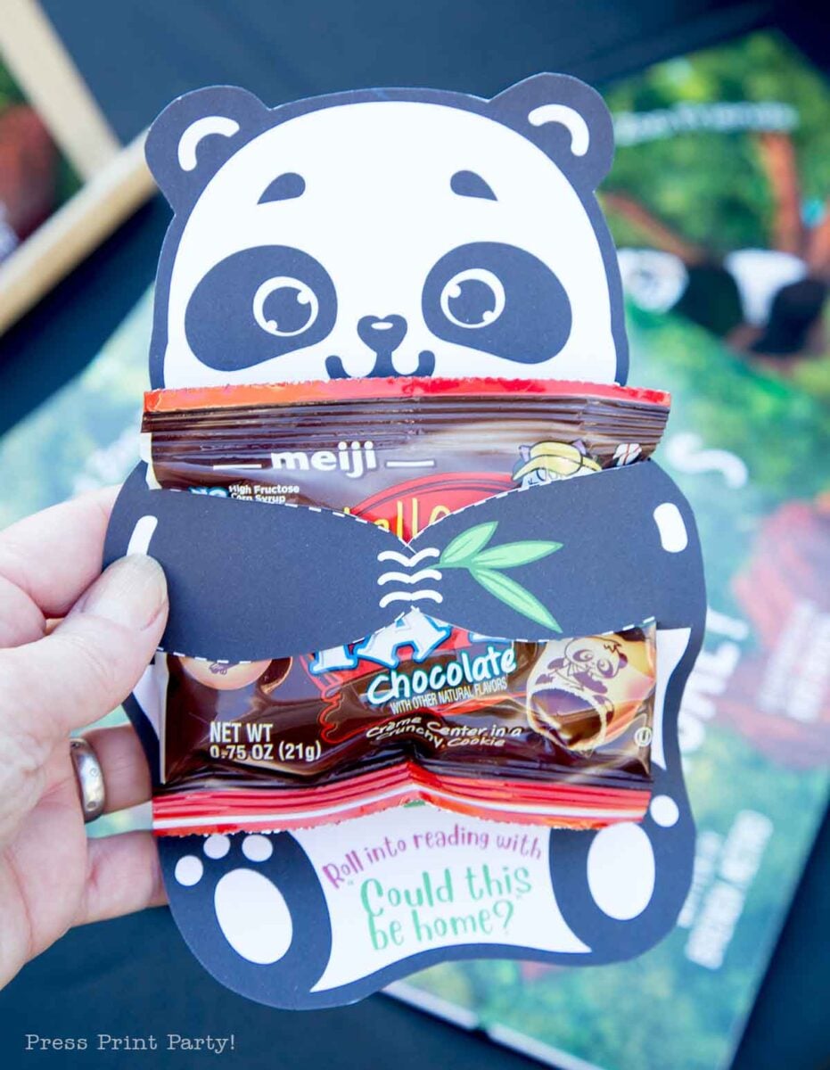 A hand holds a panda-shaped paper holder, featuring a panda with balloons, containing two Meiji chocolate candy packets. At the bottom, text reads, "Roll into reading with Could this be home?" Books are visible in the background.
