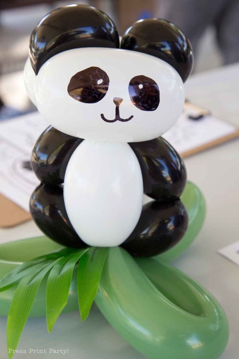 A panda with balloons made from black, white, and green latex sits on a table, with green balloon "leaves" at its base, resembling it standing on grass.