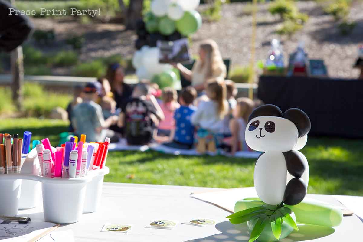 A panda with balloons figurine sits on a table with colorful markers and paper. In the background, a group of children and an adult gather on the grass outdoors for an activity.