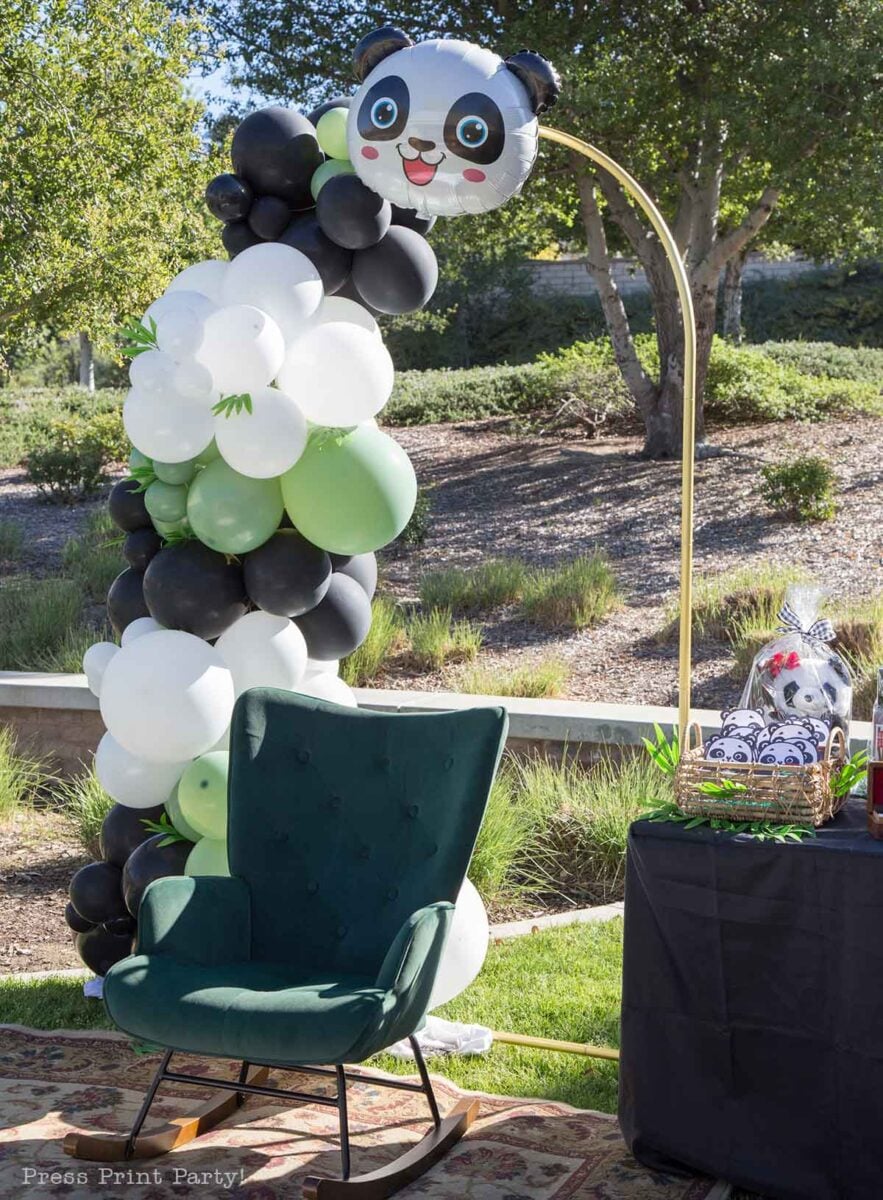 A dark green armchair sits outdoors beside a panda with balloons arch, featuring black, white, and green balloons topped with a panda face. A table displays a panda-themed gift basket, surrounded by trees and lush greenery.