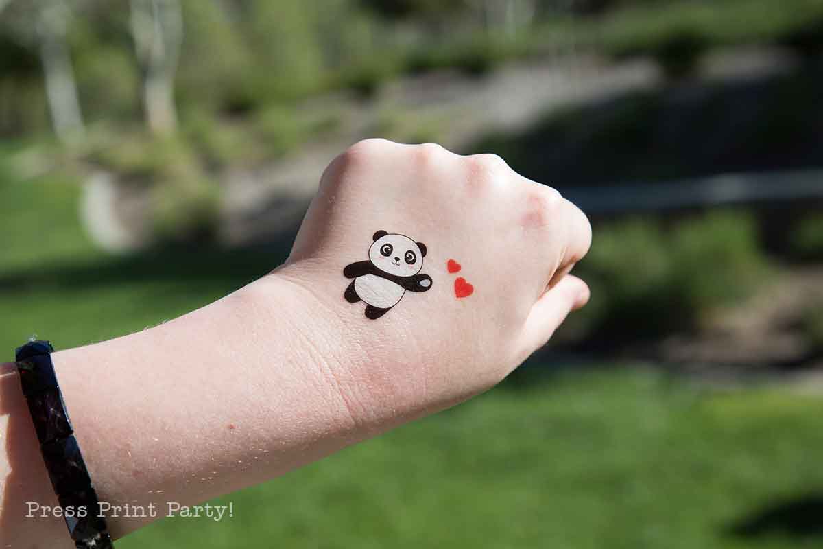 A hand with a black bracelet shows a cute panda with balloons and two red hearts as temporary tattoos on the back. The background is grassy and blurred.