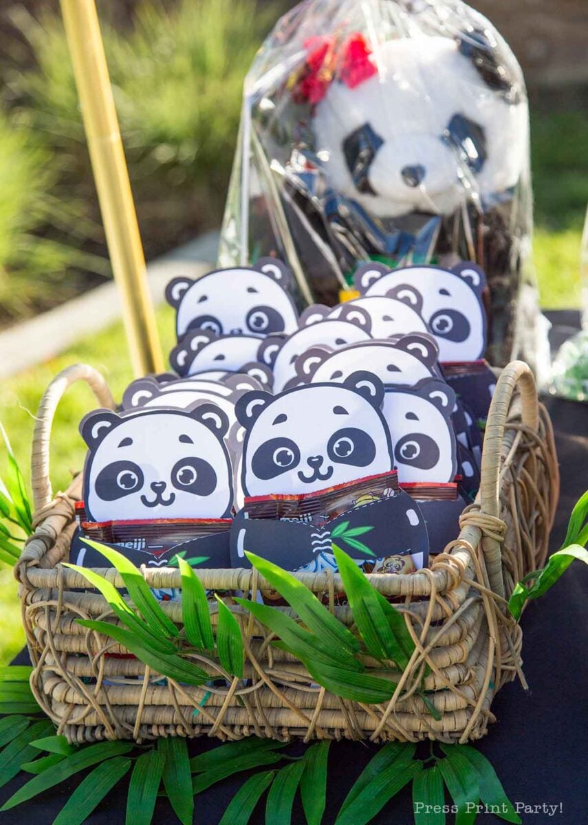 A basket filled with panda-themed party favors sits on a table decorated with green leaves, while a large panda plushie holding balloons is wrapped in plastic in the background outdoors.