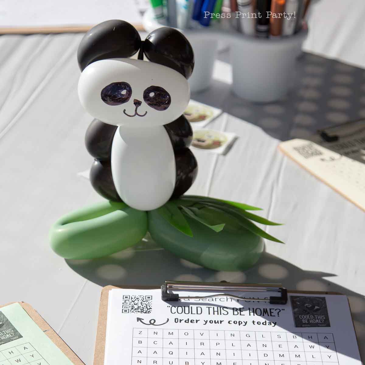 A cute panda with balloons sits on a table next to clipboards with word search puzzles and order forms, basking in the bright sunlight.