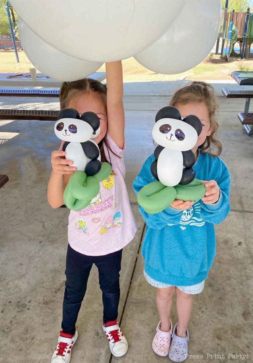 Two young children stand side by side holding balloon animals shaped like pandas in front of their faces. One child wears a pink shirt and the other a blue sweatshirt—adorable inspiration for panda party ideas under a covered outdoor area.