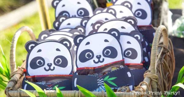 A basket filled with panda-shaped party favor bags, each featuring a cute panda face and holding treats—an adorable addition to your panda party ideas. The basket is placed outdoors with greenery in the background.