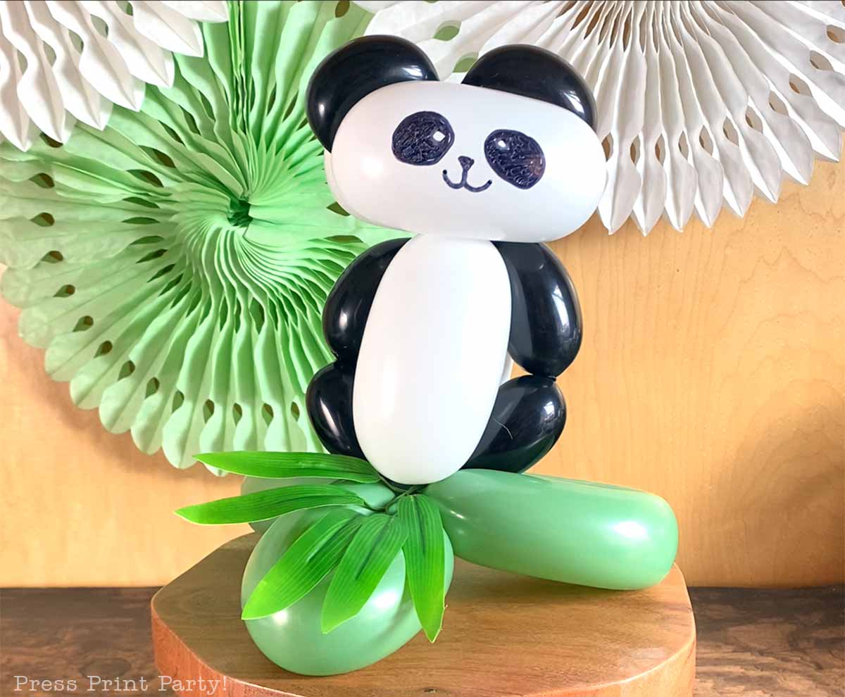 A panda with balloons made from black, white, and green balloon art sits on green balloon bamboo, with decorative paper fans in the background.