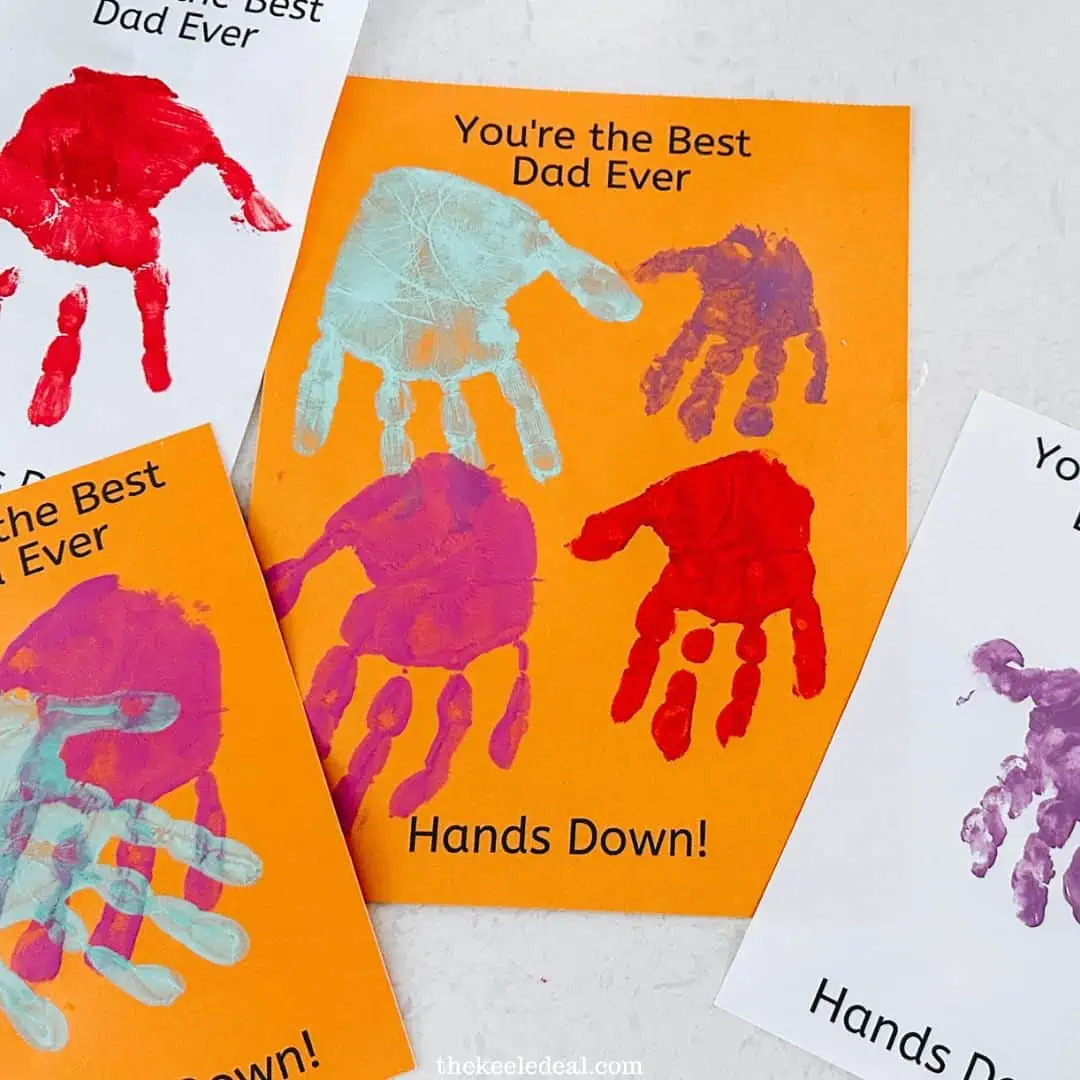 Four orange and white cards with colorful handprints and the text “You’re the Best Dad Ever. Hands Down!” are displayed on a white surface. The handprints are made with bright red, purple, and teal paint.