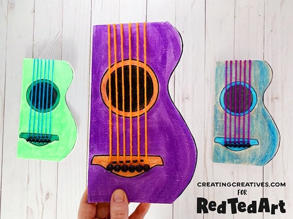 Three colorful DIY guitar crafts made from cardboard and yarn, colored green, purple, and blue, are displayed on a white wood surface. One is being held up, and the others lie flat.