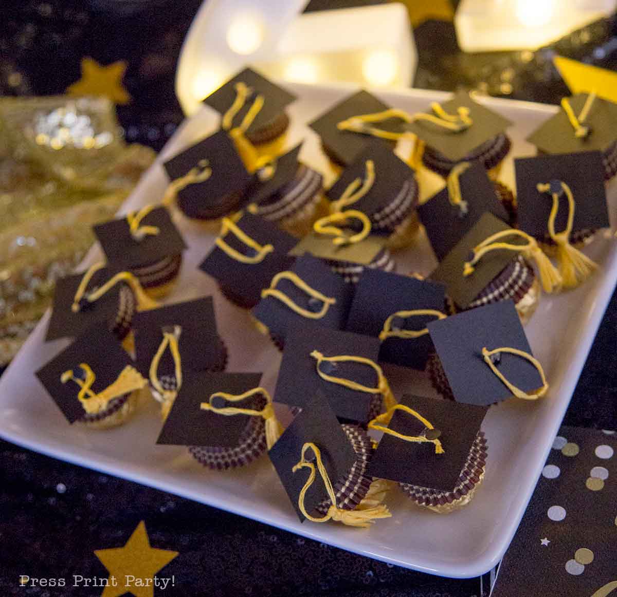 A plate of small Ferrero Rocher graduation caps, decorated with black square tops and yellow tassels, arranged on a white dish at a festive party setting.