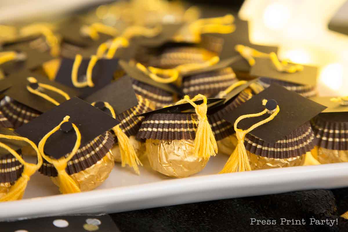 Gold-wrapped round chocolates are decorated with black paper graduation caps and yellow tassels, arranged neatly on a white tray for a graduation celebration.