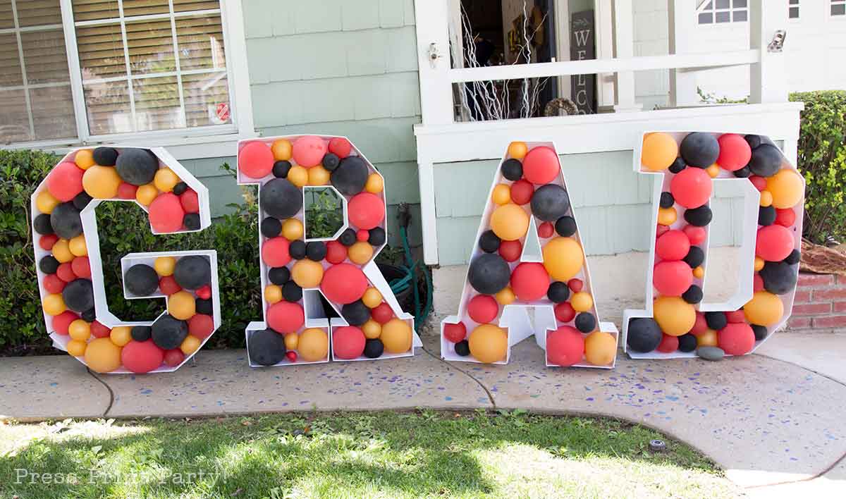 Large letters spelling "GRAD" are decorated with red, yellow, and black balloons for a theater themed graduation party. The display sits on a sidewalk in front of a house with green siding and a lawn.
