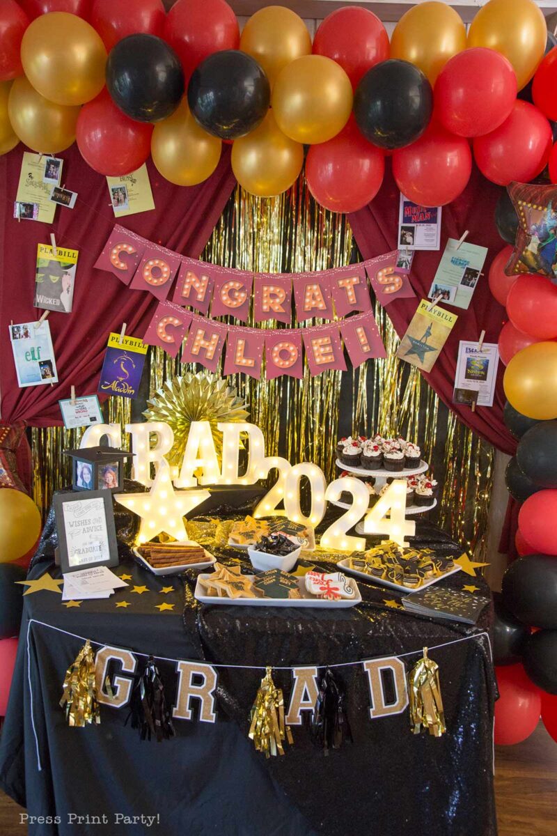 A theater themed graduation party setup with red, black, and gold balloons, a "Congrats Chloe!" banner, "GRAD 2024" lights, cupcakes, snacks, photos, and gold streamers decorating the festive table.
