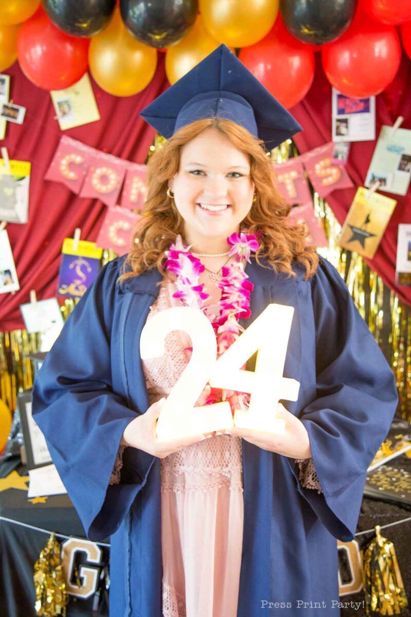 A smiling graduate in a cap and gown holds a glowing "24" sign. She wears a pink lei, standing before a theater themed graduation party backdrop with balloons, photos, and a gold "CONGRATS" banner.