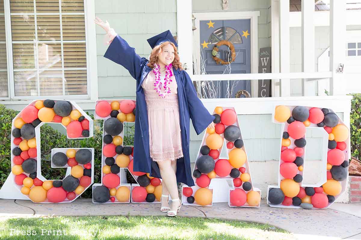 A young woman in a graduation cap and gown stands smiling with one arm raised in front of large balloon letters spelling "GRAD" at a theater themed graduation party outside a house.