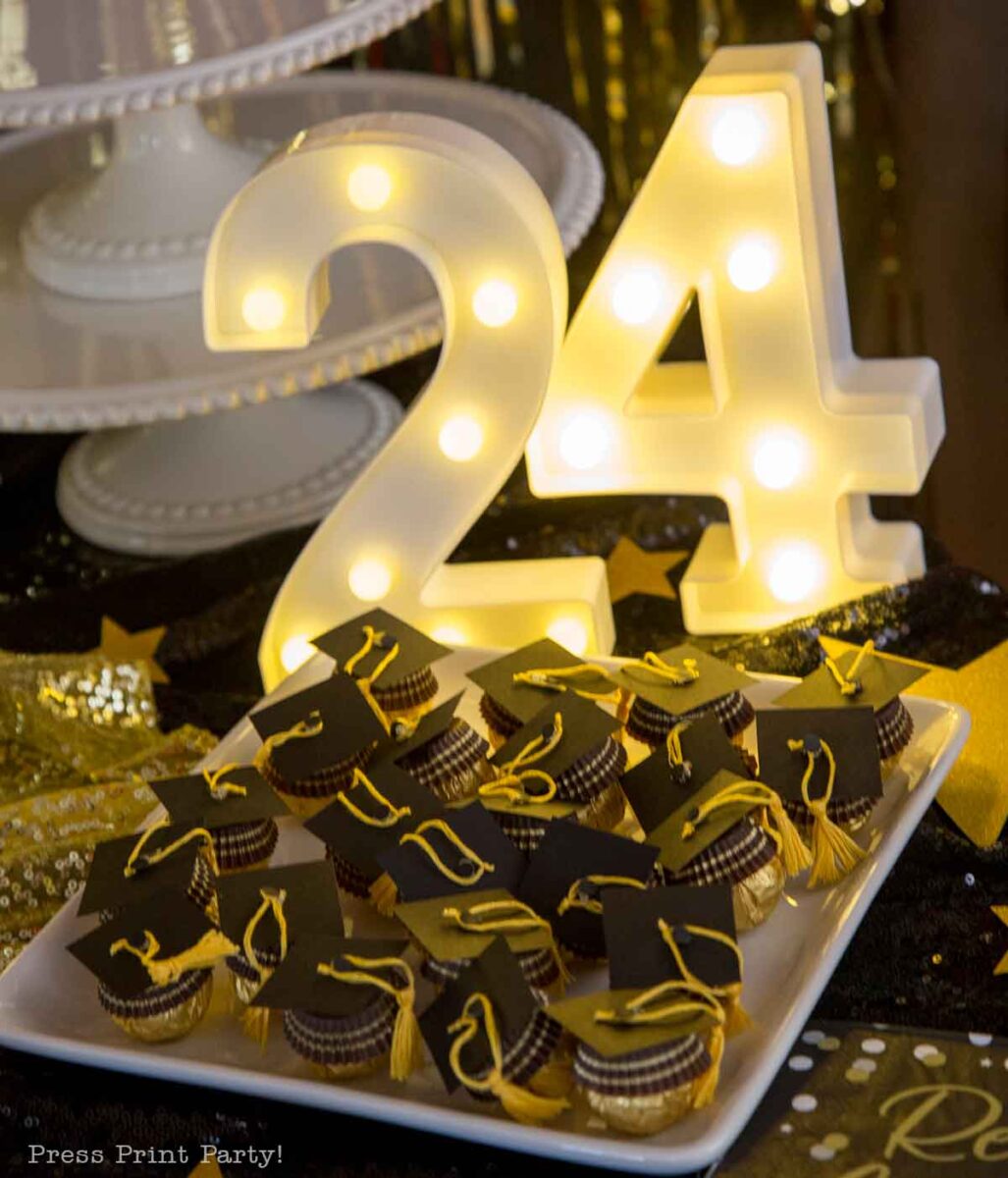 A light-up "24" sign behind a plate of mini desserts decorated with black mortarboard graduation caps and yellow tassels, set on a black and gold festive table for a theater themed graduation party.