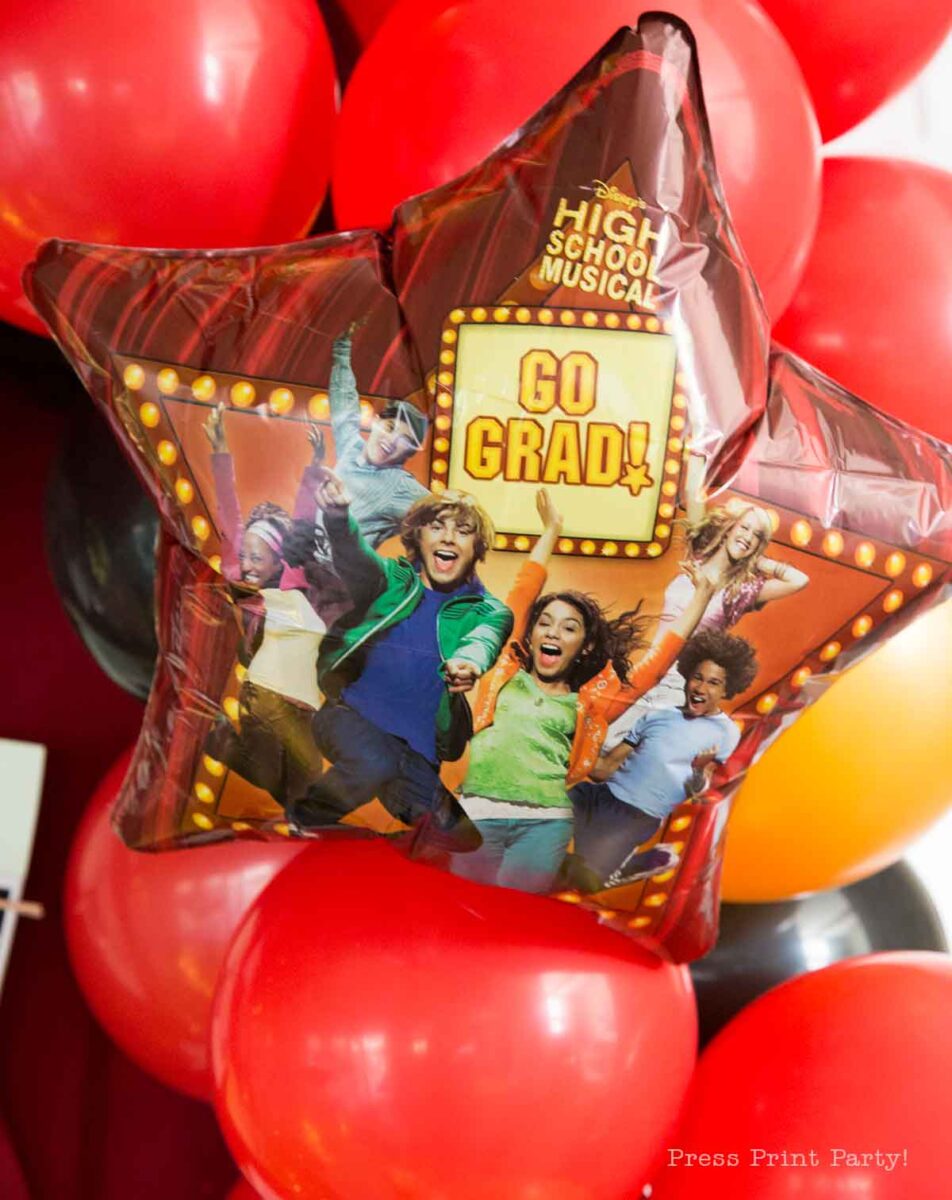 A star-shaped balloon featuring characters from High School Musical and the words "GO GRAD!" is displayed in front of red and black balloons, perfect for a theater themed graduation party.