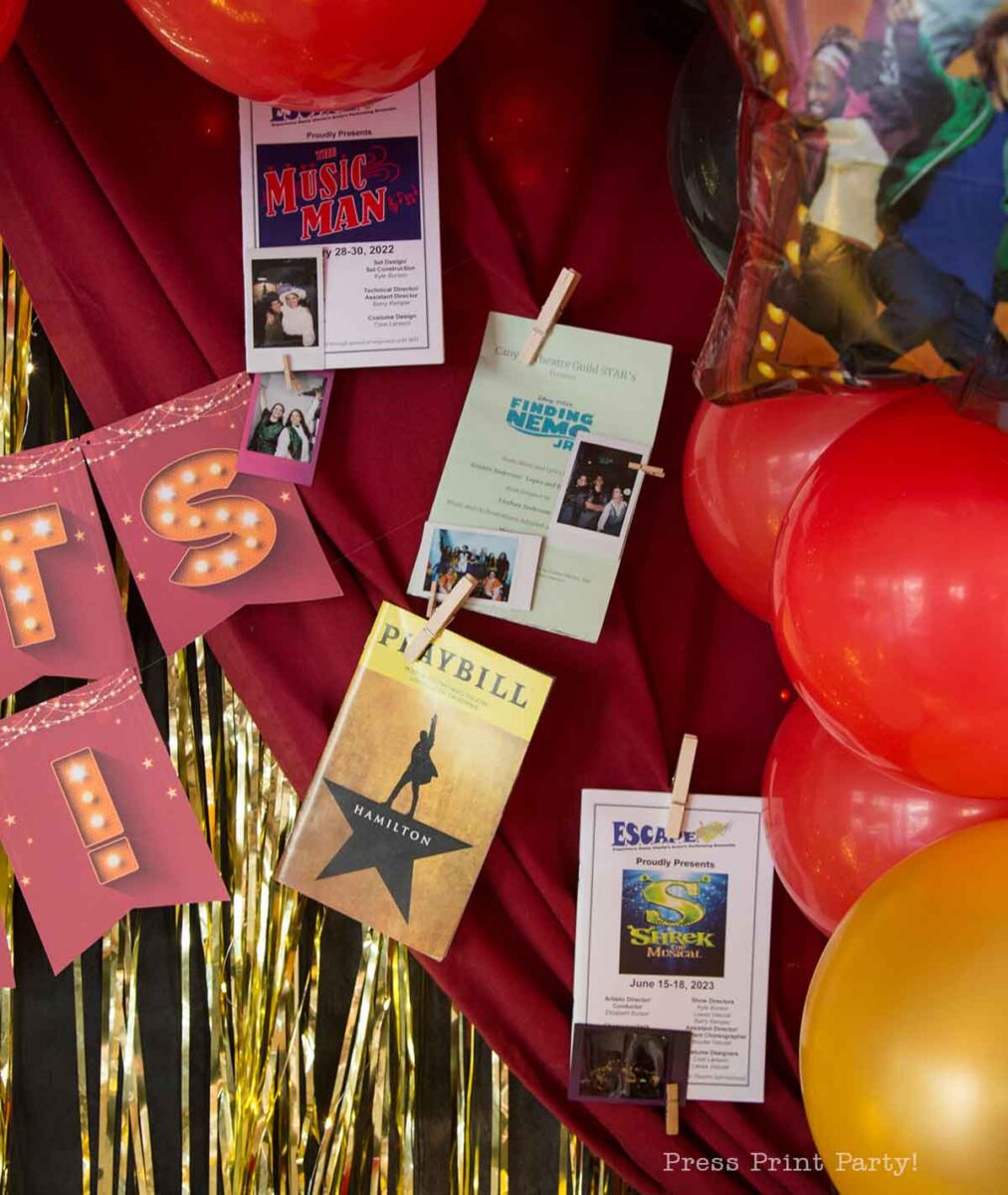 A theater themed graduation party table decorated with red and gold balloons, gold fringe, and a red cloth displays musical programs, including Playbills for βHamilton,β βShrek The Musical,β βThe Music Man,β and βHonk Jr.,β plus a pink βITS!β banner.