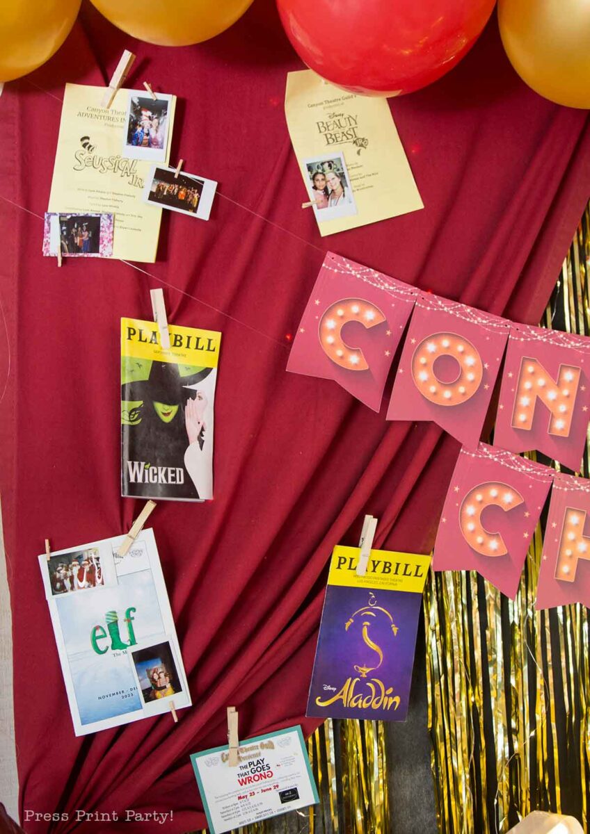 A theater themed graduation party setup features a red drape with gold streamers and balloons, decorated with Playbills, show flyers, and photos attached by clothespins, plus a pink "CONGRATS" banner in gold letters.