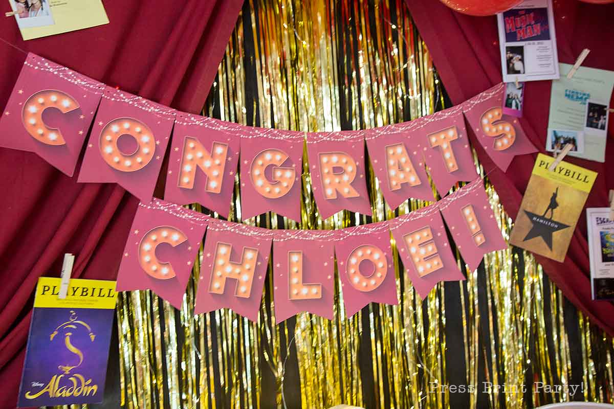 A theater themed graduation party backdrop with pink banners reading "CONGRATS CHLOE!" in marquee-style letters, surrounded by red curtains, gold tinsel, and Broadway Playbills including "Aladdin.