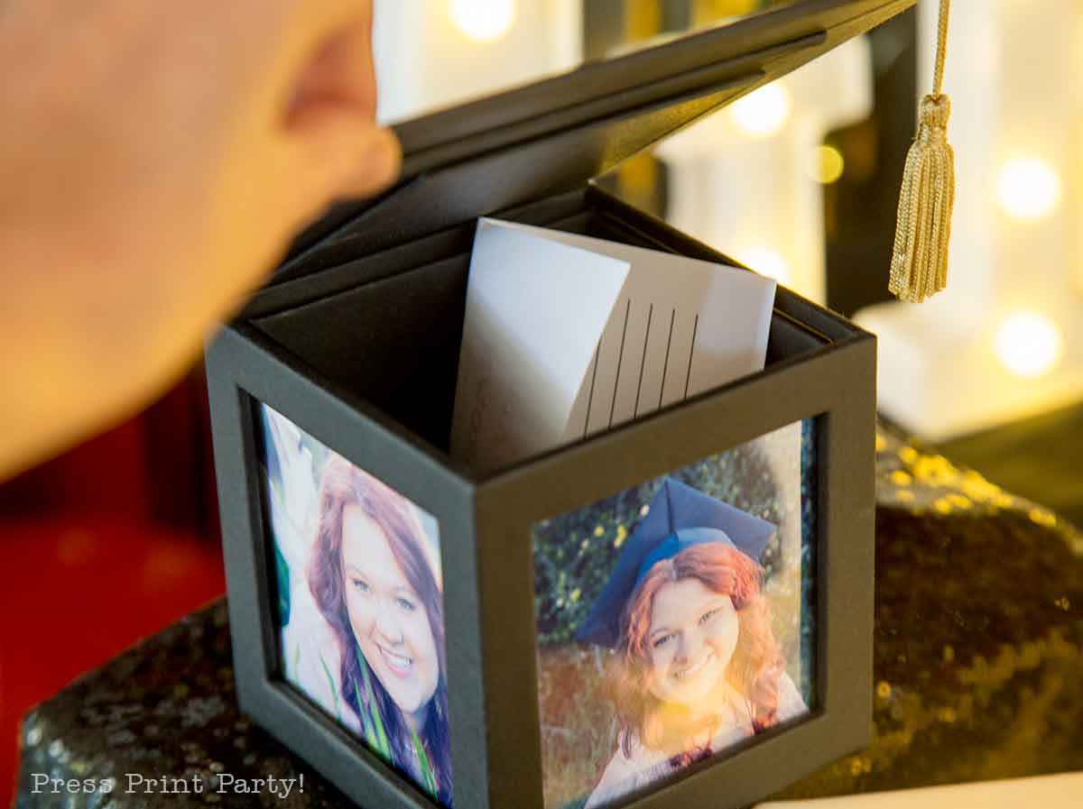 A hand opens a black box decorated with photographs of a smiling young woman at her theater themed graduation party. Inside the box are blank cards, and a gold tassel decorates the lid as warm lights glow softly in the background.