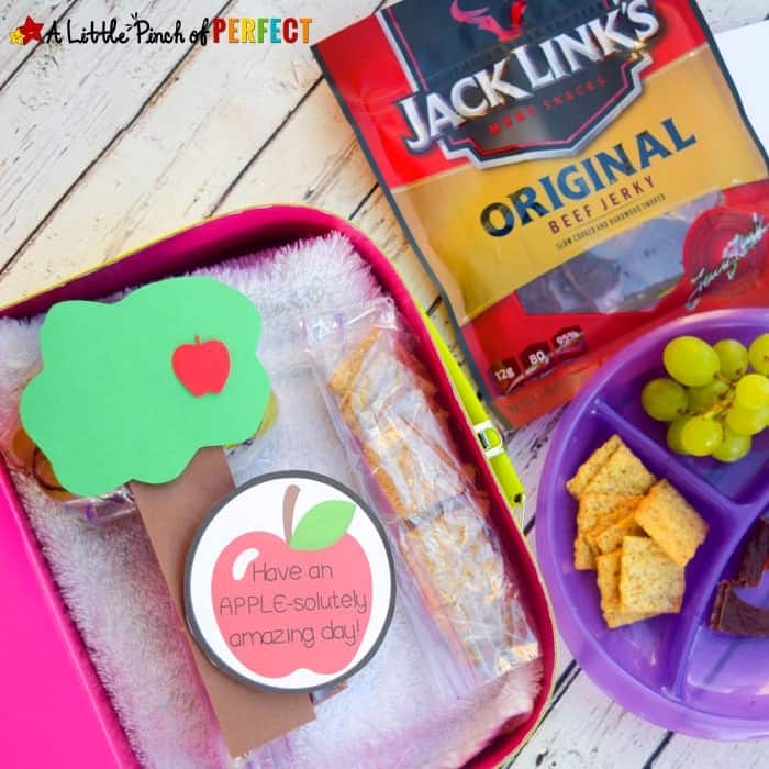 A lunchbox with a sandwich wrapped as a tree, apple-themed note, grapes, crackers, and a pack of Jack Link’s beef jerky on a wooden surface.