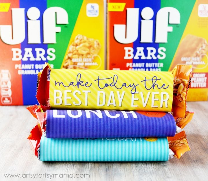 Three Jif Bars are stacked, each with a brightly colored wrapper. The top says “make today the BEST DAY EVER,” the middle says “LUNCH,” and the bottom is partially visible. Jif Bars boxes are in the background.