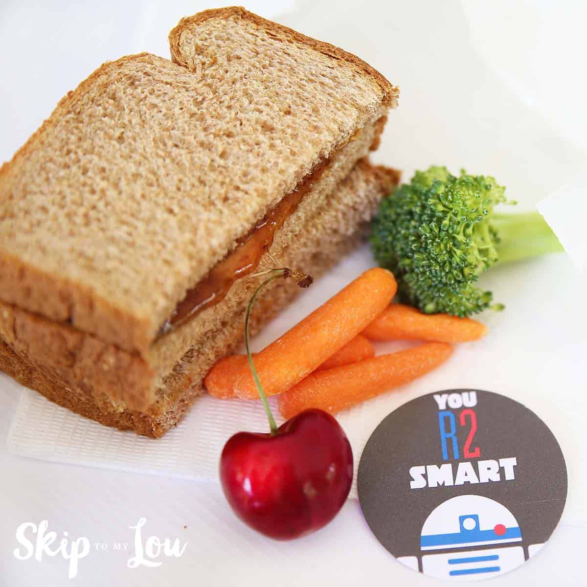 A peanut butter and jelly sandwich with baby carrots, a cherry, broccoli, and a round card that says “You R2 Smart” with a small graphic of R2-D2.