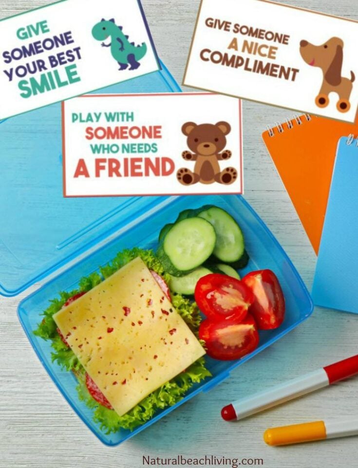 A lunchbox with a sandwich, cucumbers, and tomatoes sits next to notebooks and a pen. Three cards read: “Give someone your best smile,” “Give someone a nice compliment,” and “Play with someone who needs a friend.”.