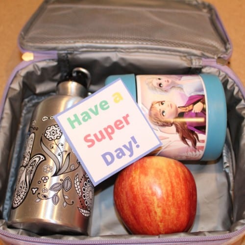 Open lunchbox with a silver water bottle, a Frozen-themed container, a red apple, and a note reading “Have a Super Day!”.