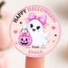A finger holds one of our 6 Pink Coquette Halloween Ghost Round Labels, featuring a cute ghost with a pink bow holding a pink pumpkin and the message “Happy Halloween! from EMMA.” The light background displays blurred stars and pink pumpkins, making these labels perfect for adding a sweet touch to your gifts.