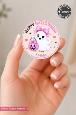A hand holds one of the 6 Pink Coquette Halloween Ghost Round Labels, a round pink sticker featuring an adorable ghost with a pink bow and blush, a pumpkin, and the text “Happy Halloween! from Emma.” The sticker also includes Canva and “Press Print Party!” logos.