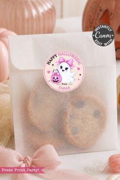 A translucent treat bag filled with cookies is sealed with a round sticker from the 6 Pink Coquette Halloween Ghost Round Labels set, featuring a cute ghost with a pink bow and pumpkin. The charming labels add a festive touch, while pink decorations and pumpkins surround the bag.
