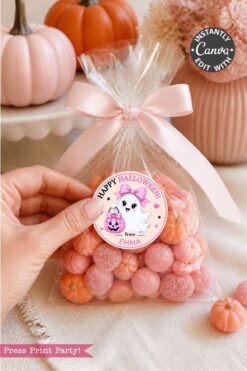 A hand holds a clear bag of pink candies tied with a ribbon. The bag is decorated with one of the 6 Pink Coquette Halloween Ghost Round Labels, featuring a “Happy Halloween” sticker with a ghost and a pink pumpkin, and “from Emma” written below. Pink pumpkins and decor are in the background.