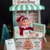 A festive “North Pole Cookie Stand” display featuring an elf doll, cookie signs, a holiday menu, and cheerful red, green, and white Christmas decor. Adorned with cookies and a gingerbread graphic, this charming setup becomes even more delightful when paired with the 6 Pink Coquette Halloween Ghost Round Labels for a whimsical touch to your holiday celebrations.