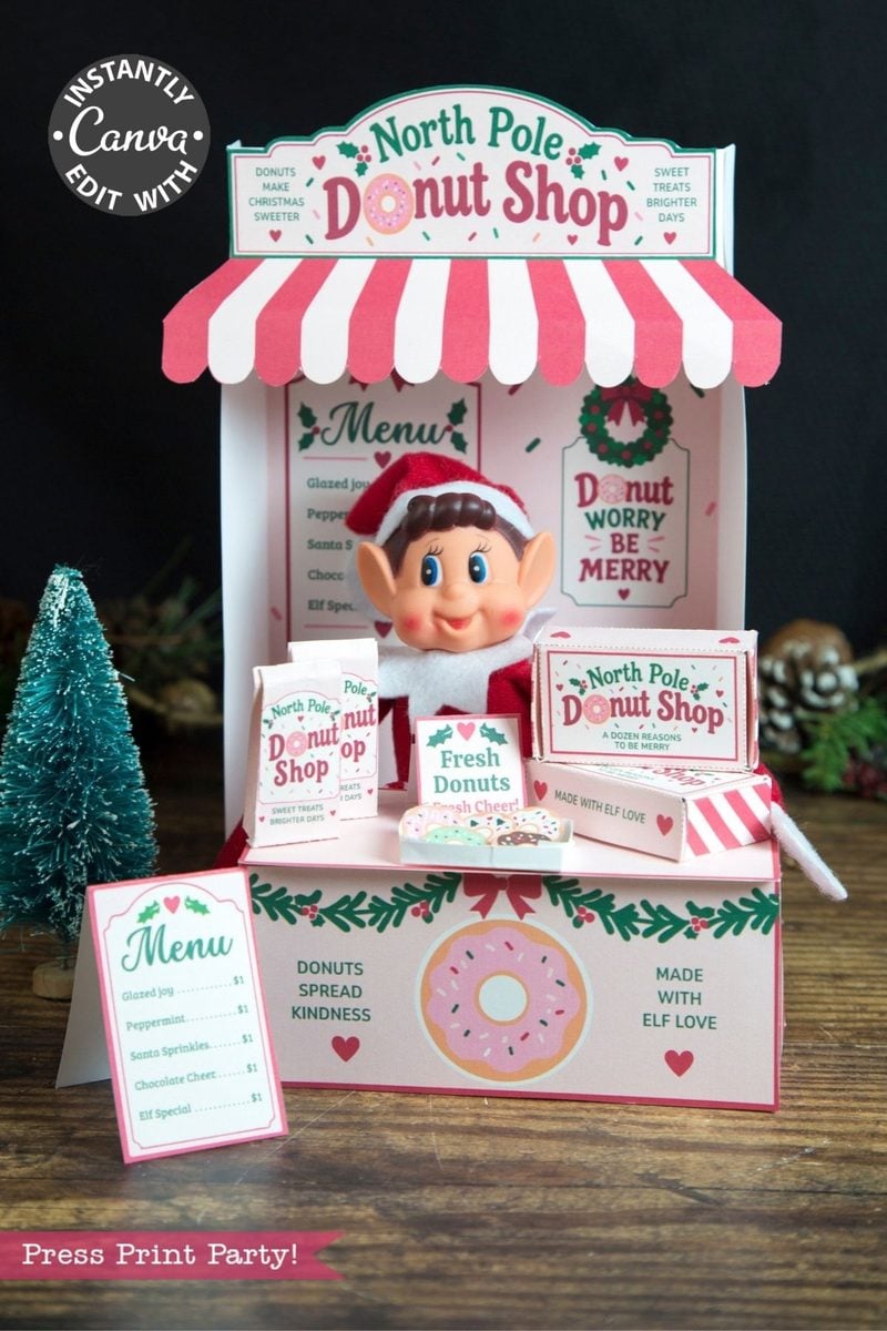 A festive North Pole Donut Shop display features an elf doll, donut menus, pink and green holiday decor, and mini donut boxes adorned with 6 Pink Coquette Halloween Ghost Round Labels, with a small tree beside the stand.