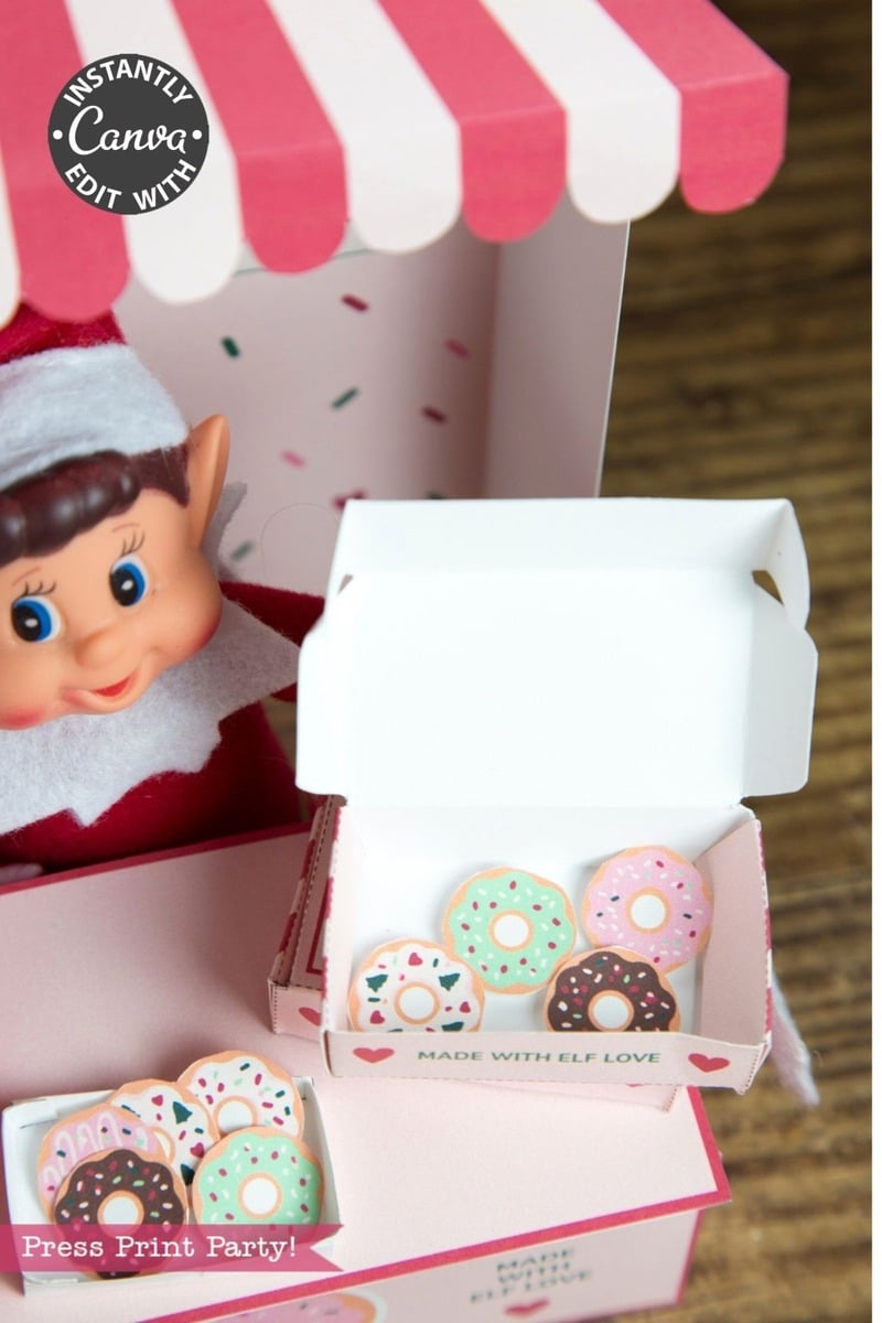 An elf doll sits in a Pink Coquette-inspired booth beside two open boxes of colorful mini frosted donuts with sprinkles. The boxes, each adorned with a charming Pink Coquette Halloween Ghost Round Label from the “6 Pink Coquette Halloween Ghost Round Labels” set that reads “MADE WITH ELF LOVE,” rest on the wooden surface below the striped awning.
