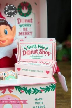 A smiling elf doll sits beside pastel pink boxes labeled “North Pole Donut Shop,” decorated with festive designs, donuts, and holiday messages like “Donut worry be merry” and “Made with elf love.” The scene is adorned with 6 Pink Coquette Halloween Ghost Round Labels, blending charming whimsy and cheerful holiday spirit.