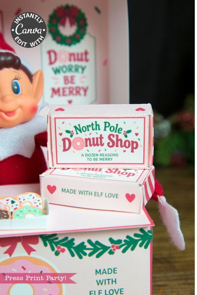 A smiling elf doll sits beside pastel pink boxes labeled “North Pole Donut Shop,” decorated with festive designs, donuts, and holiday messages like “Donut worry be merry” and “Made with elf love.” The scene is adorned with 6 Pink Coquette Halloween Ghost Round Labels, blending charming whimsy and cheerful holiday spirit.